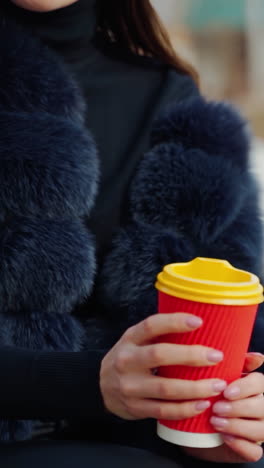 female hands hold a cup of beverage outdoors. beautiful woman in a fur coat sitting in the park and drinking from plastic glass. slow motion. vertical video
