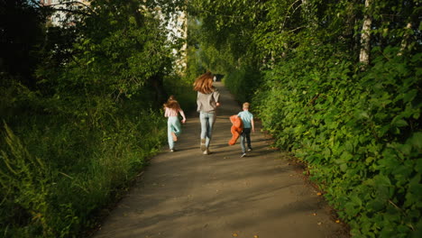 rear view of energetic woman running down shaded path with her children, boy gripping teddy bear while sister sprints ahead, path lined with dense greenery under warm natural light
