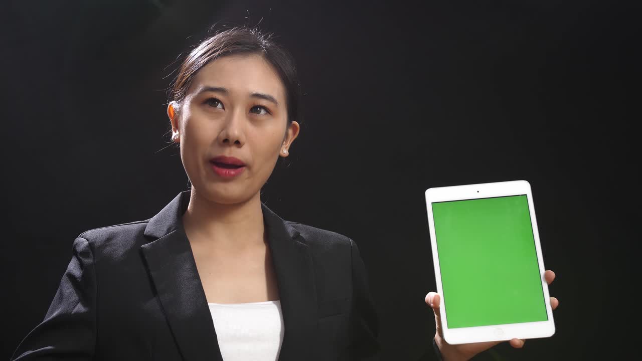 블랙 스크린 스튜디오에서 말하는 동안 녹색 스크린 태블릿을 들고 가리키는 비즈니스 슈트를 입은 아시아 스피커 여성의 클로즈업