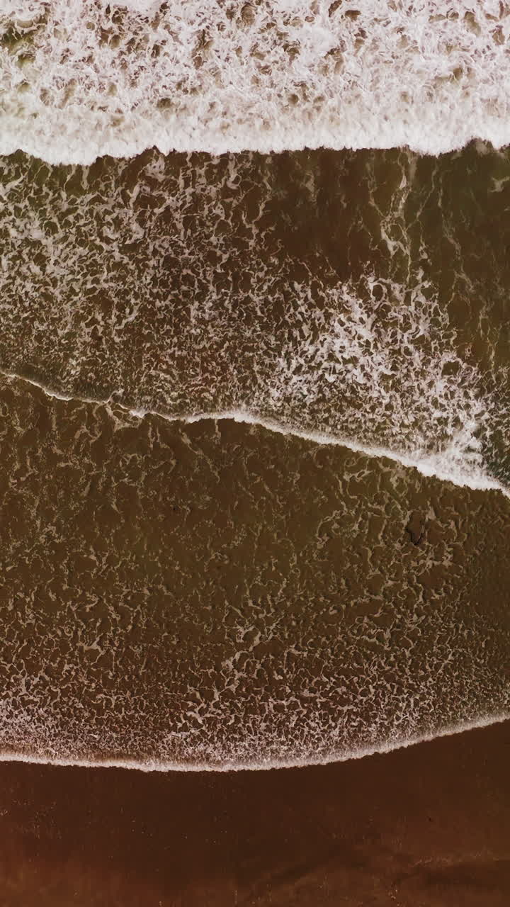 White broad foamy waves covering the sandy beach. Drone footage descending over the ocean coast on windy weather. Vertical video
