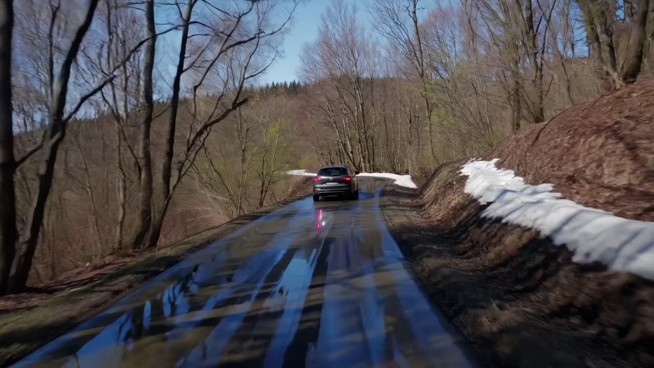 A rear-view video captures a car driving on a wet, snow-lined forest road