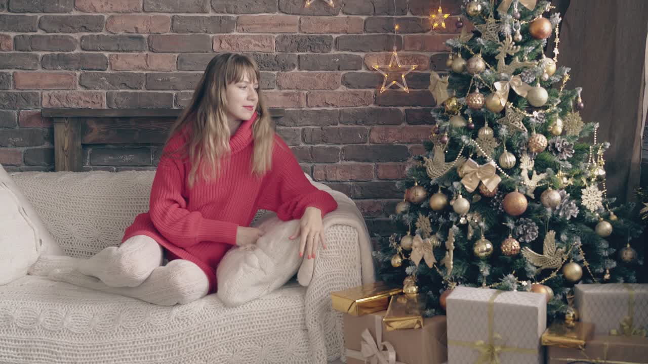 una joven rubia con suéter rojo se sienta cerca del árbol de navidad