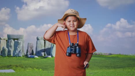asian boy with a hat and binoculars using the magnifying glass then making call me gesture while traveling in stonehenge. boy researcher examines something, travel tourism adventure concept