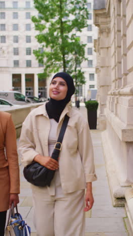 Vertical-Video-Of-Two-Muslim-Businesswomen-Wearing-Hijabs-With-Modern-Business-Suits-Walking-Along-Pavement-To-Work-Past-City-Office-Buildings