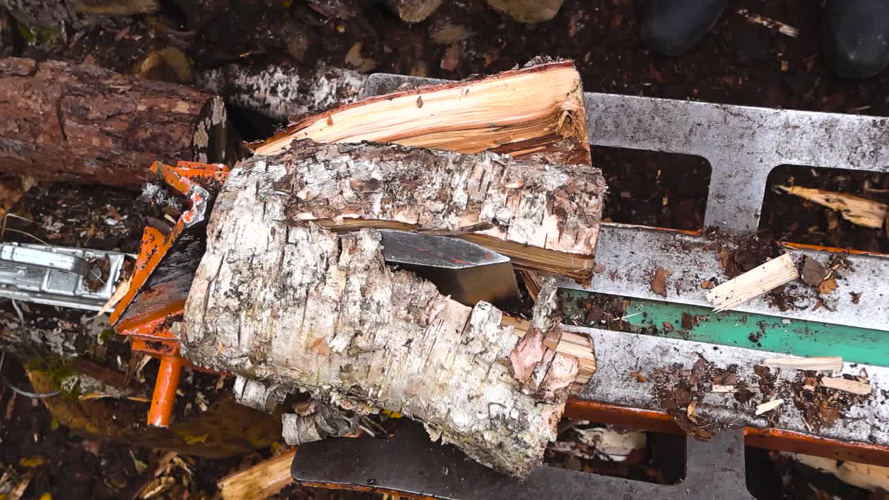 Top down footage of a birch tree log or timber being split in half and to pieces by a wood splitter machine in slow motion. Sawdust and wood chips flying around on the damp brown colored soil below