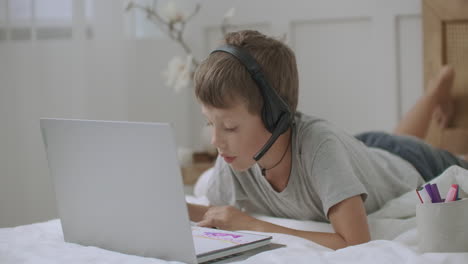 funny school boy is listening teacher in headphones during remote learning session and drawing picture in copybook