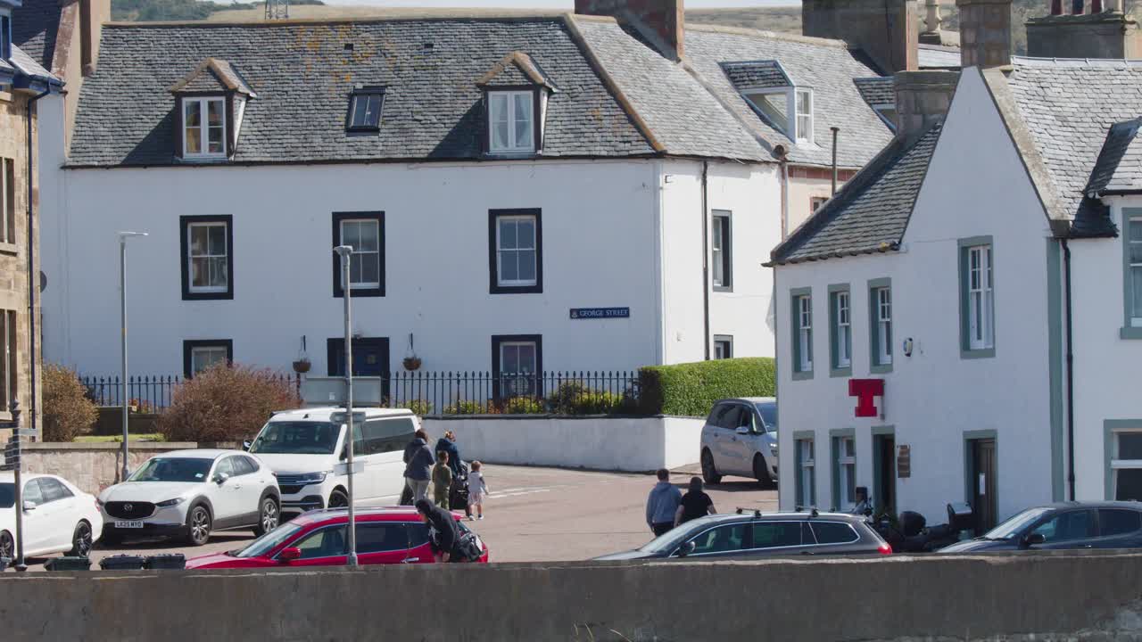 People walk along a sunny street lined with historic stone buildings and parked cars