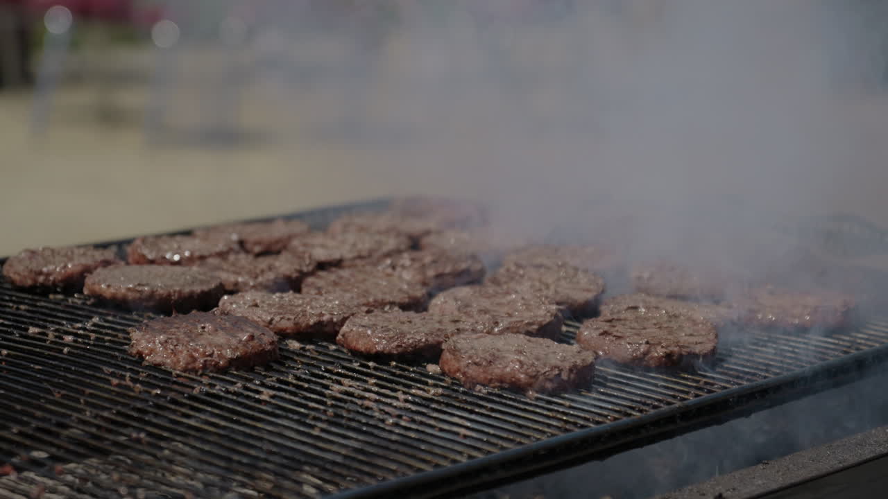 Inquadratura ampia di hamburger su un barbecue a carbone fumante e unto, su una griglia commerciale, in un pomeriggio estivo, al rallentatore