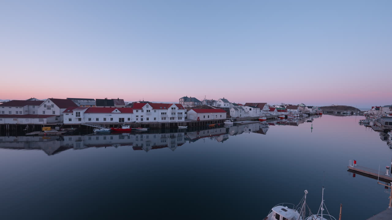 Sonnenaufgang in Henningsvær, Lofoten, Norwegen, mit dem Fischerdorf, Häusern am Wasser, einem rosa-blauen Himmel, Möwen und spiegelglattem Wasser, das den Himmel reflektiert.