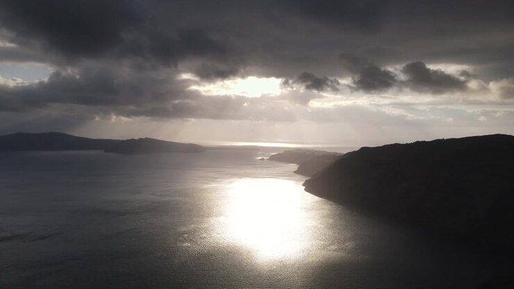 Aerial drone footage captures the volcanic caldera of Santorini with the Aegean Sea reflecting sunlight through heavy storm clouds. Smooth lateral drone movement reveals the dramatic coastline.