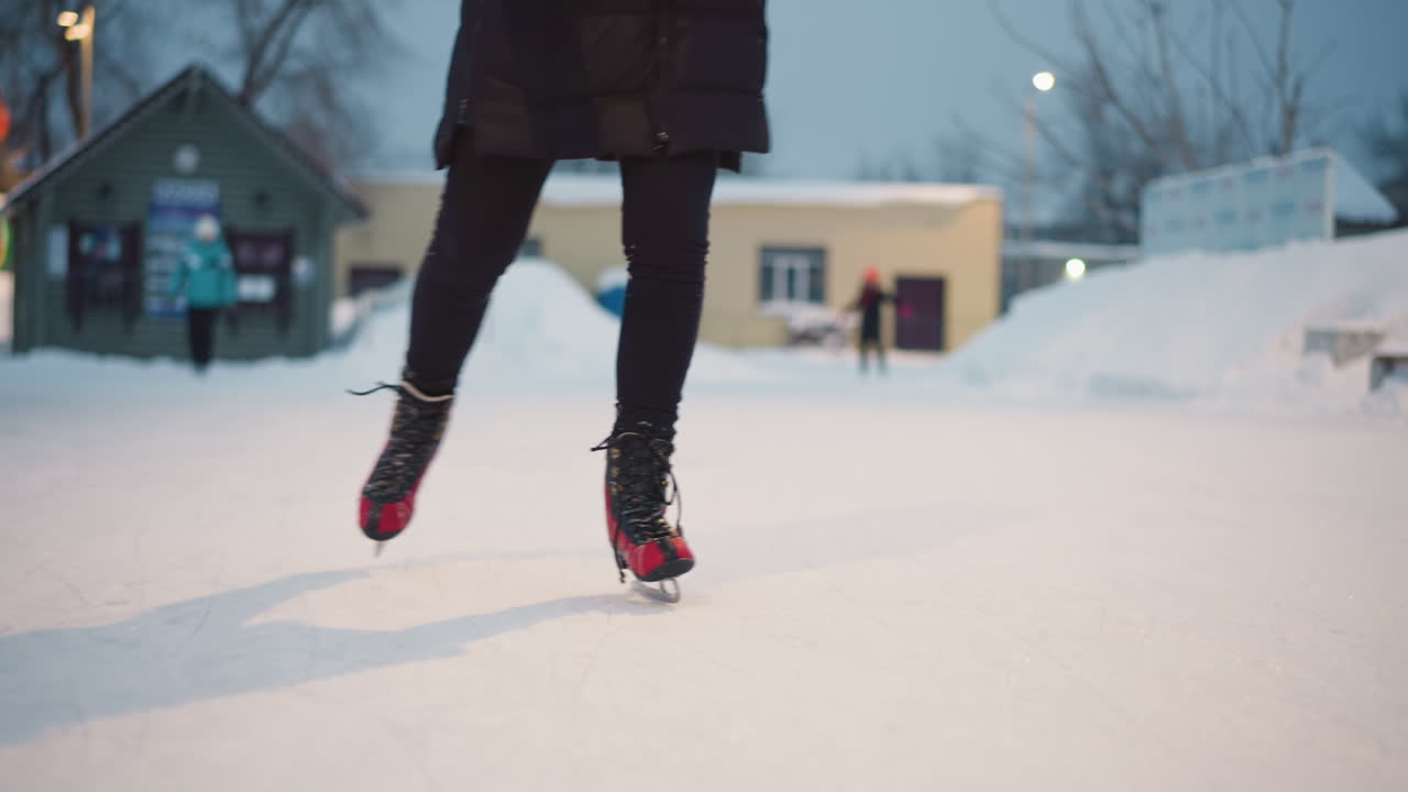 Person skating on outdoor ice rink in red skates during winter evening, snow piled around frozen surface, motion captured with dynamic energy, reflecting seasonal recreation,fitness, leisure atmosphere
