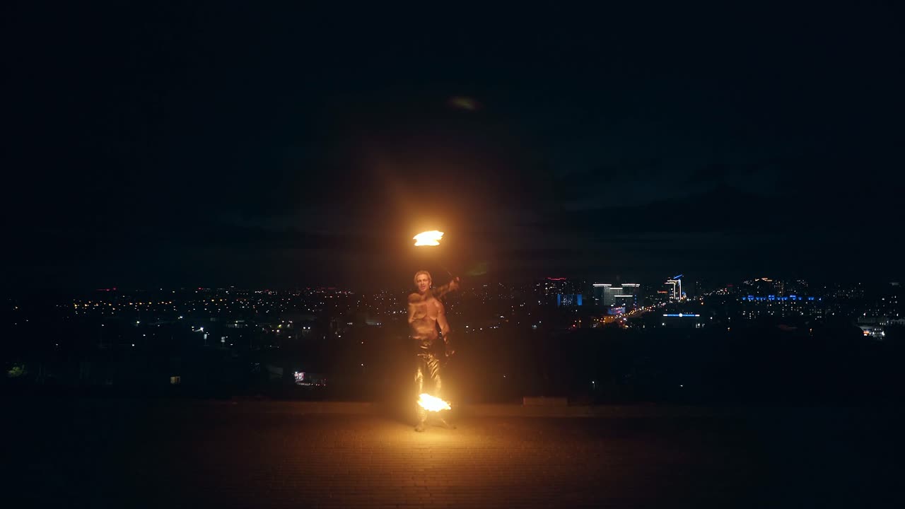 Young blond male does fire dance with two torches spins torches at night on black background
