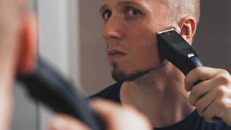 guy-shaves-beard-with-trimmer-looking-in-mirror-closeup