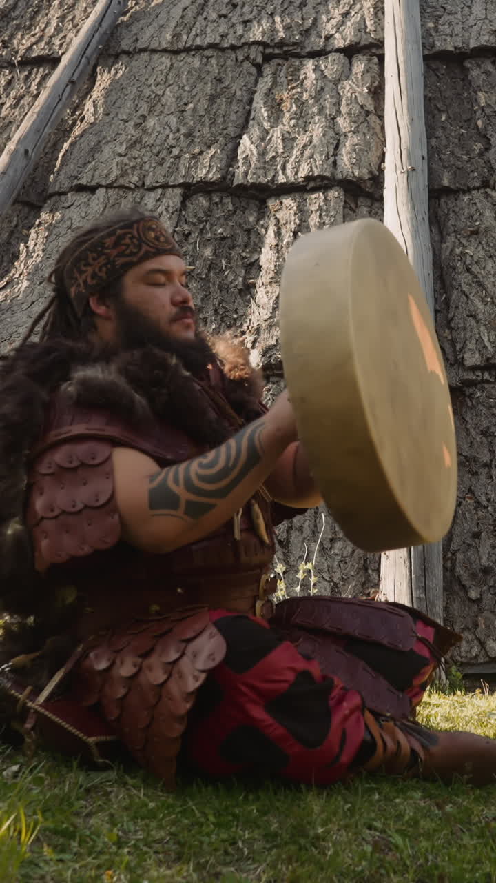 Obese man in leather clothes with fur repairs shamanic drum sitting near old ail. Musician prepares instrument for performance. Spiritual fest