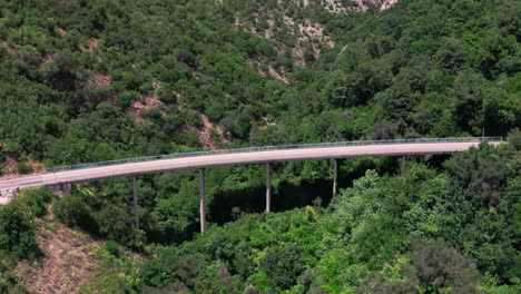 A-curving-beam-bridge-over-a-steep-mountain-ridge-covered-with-trees-and-bushes-near-Milocher,Montenegro,with-a-white-car-and-a-white-van-passing-by-on-a-sunny-summer-day,-aerial-shot