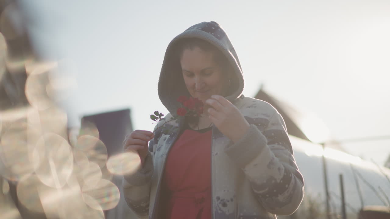 Donna bianca nel giardino sul retro all'alba che tiene fiori rossi in un maglione con cappuccio, silhouette e bagliore solare con bokeh morbido, umore riflessivo e nostalgico, case sullo sfondo, lento momento domestico contemplativo.