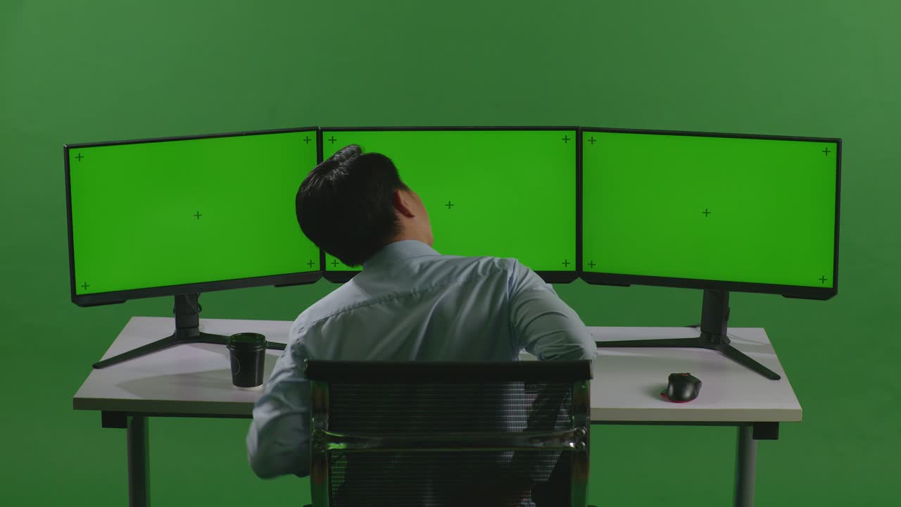 녹색 스크린 스튜디오에서 모의 다중 컴퓨터 모니터로 작업하는 동안 허리 통증이 있는 아시아 남자의 뒷면