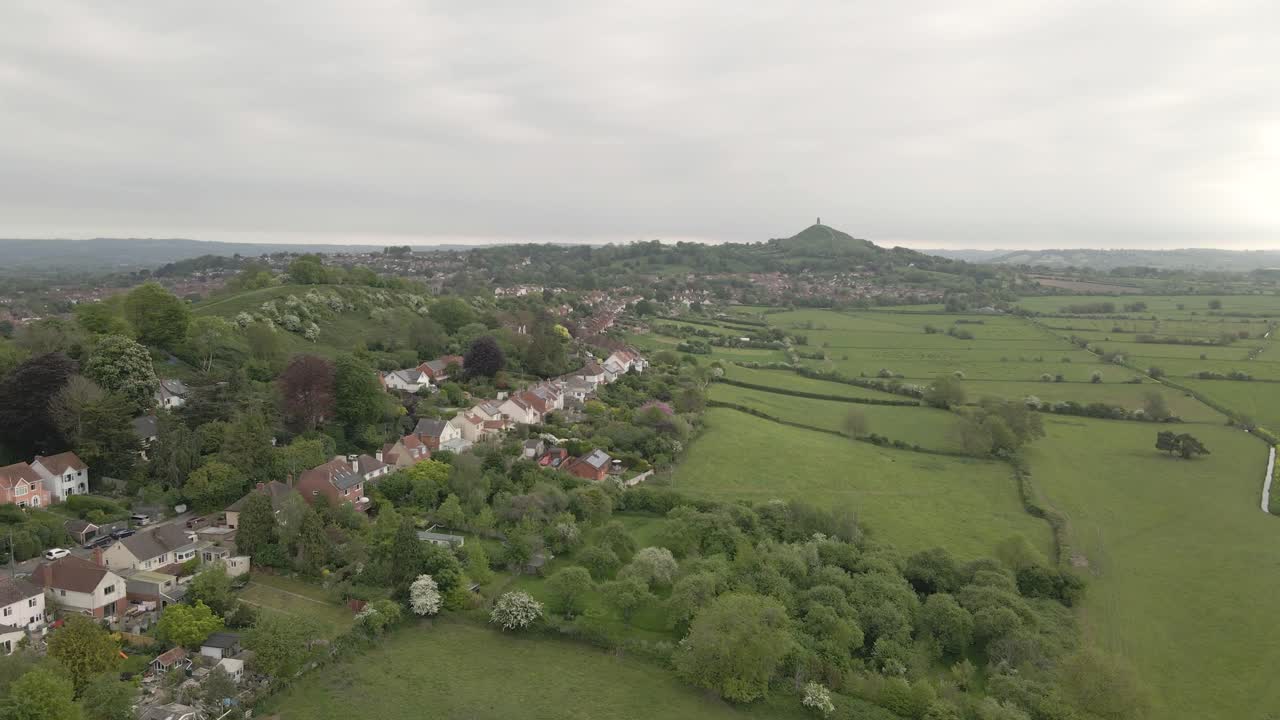 Aerial view of Glastonbury, drone rotating to the left over the houses and the Main Street leading to the Tor. green fields around the houses . 4K, 60fps.