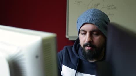 young bearded man with hat sit back on his chair in front of computers talking with colleagues, meanwhile looking on monitor