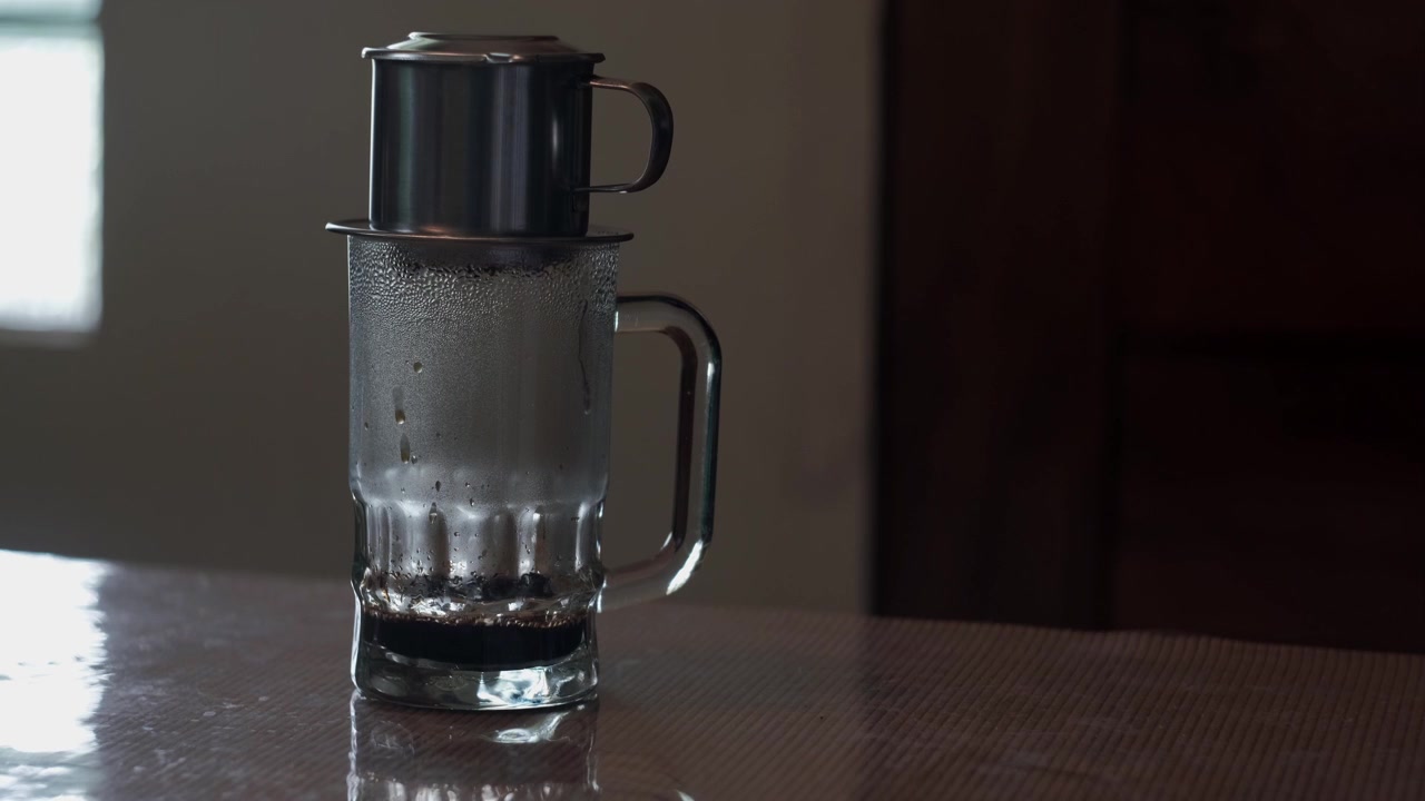 Statische Aufnahme der traditionellen vietnamesischen Phin-Kaffeezubereitung in Innenräumen, mit langsamen Kaffeetropfen, die in einen klaren Glasbecher auf einer Tischplatte fallen.