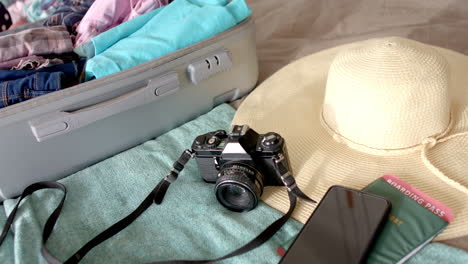 a suitcase lies open, filled with clothes, alongside camera, hat, and phone