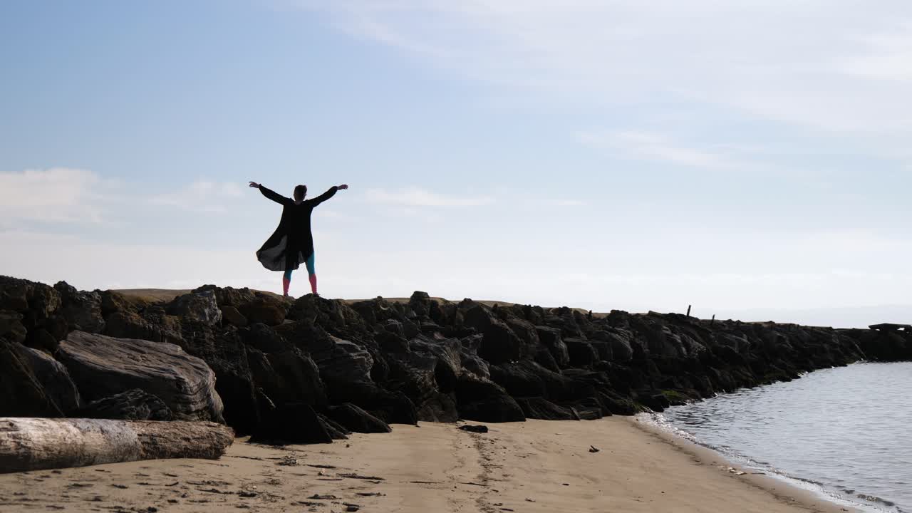 Woman stands and stretches on rocky pier by the beach on a sunny day