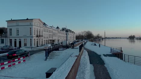 Sideways-sunset-aerial-movement-revealing-the-cityscape-facades-of-Dutch-Hanseatic-medieval-tower-town-Zutphen-in-The-Netherlands-with-snow-on-the-boulevard-in-the-foreground-along-the-river-IJssel