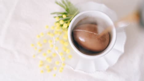 Draufsicht-Auf-Heißen-Kaffee,-Der-Auf-Ein-Braun-Bemaltes-Osterei-In-Einer-Weißen-Tasse-Gegossen-Wird-Und-Es-Auf-Weißem-Tischhintergrund-Zerbricht