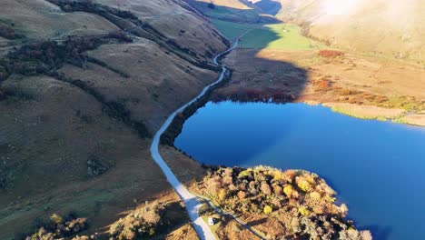 Aerial view of a winding road beside a serene lake, surrounded by mountains, captured during sunrise