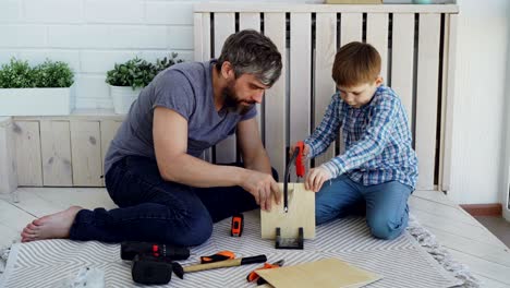 el hijo útil está aserrando una hoja de madera con la sierra de mano mientras su padre está midiendo el corte con el carrete de medida y hablando con su hijo. construcción, familia unida y concepto de la infancia.