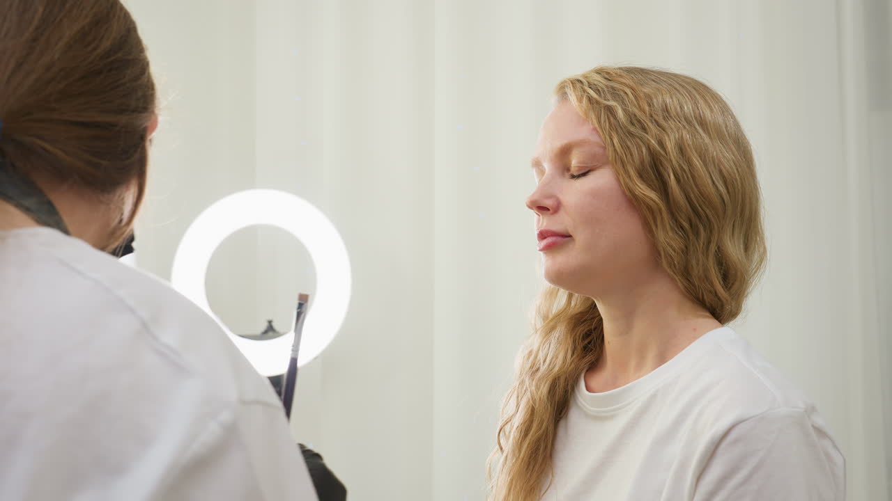 Beautician wearing black gloves and mask applies cream to client eyebrow with brush while gently shifting client s hair aside. Client with blonde wavy hair sits calmly in bright, clean beauty salon
