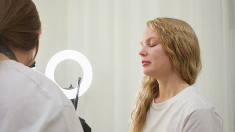 beautician wearing black gloves and mask applies cream to client eyebrow with brush while gently shifting client s hair aside. client with blonde wavy hair sits calmly in bright, clean beauty salon