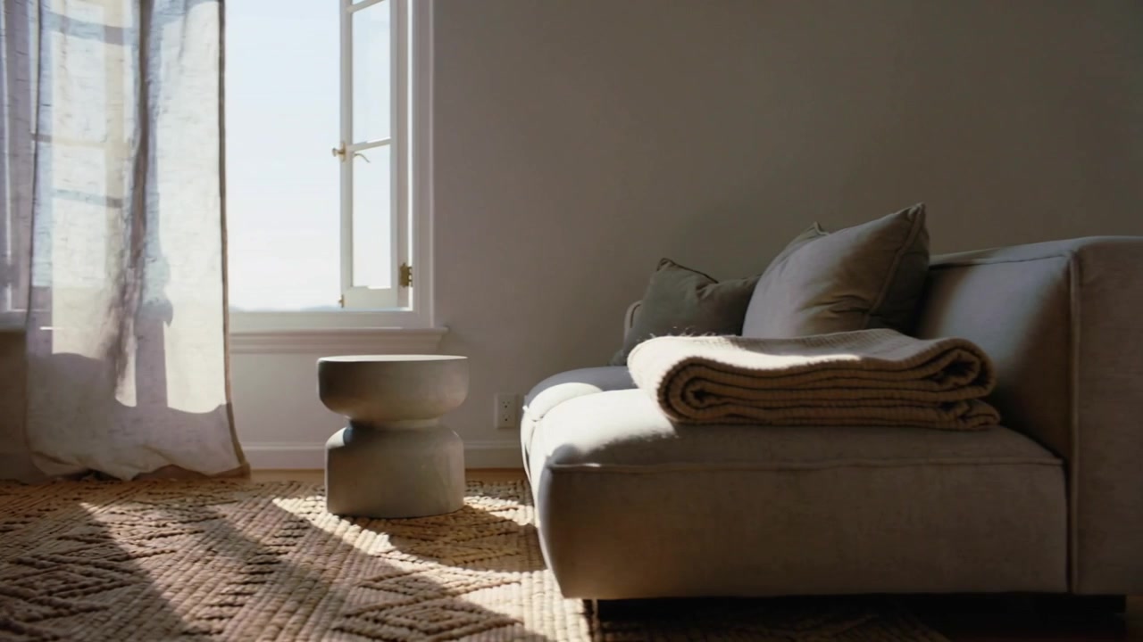 Tenda trasparente che si solleva mentre la telecamera si sposta verso una chaise longue in casa, lasciando che la luce del sole mostri il tappeto