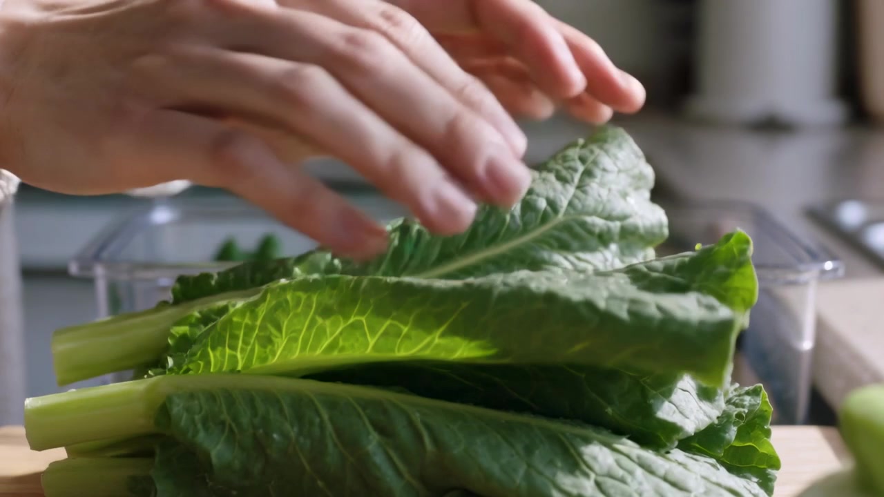 Mani con un maglione beige che mettono verdure in un contenitore trasparente, sistemando i gambi sul bancone della cucina per la conservazione.
