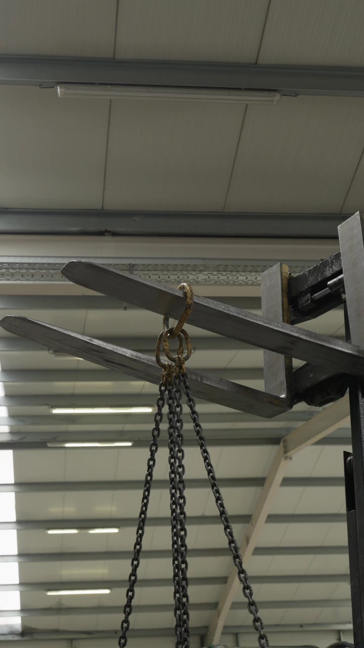 Forche del carrello elevatore sollevate con catene di sollevamento appese a un gancio all'interno di un magazzino industriale.