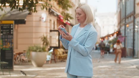 caucasian mature businesswoman in suit using mobile gps to find a direction walking on city street