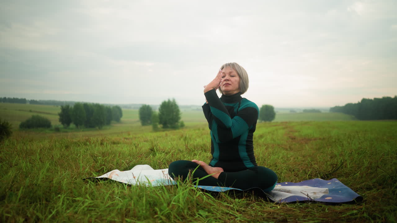 Woman seated on yoga mat in misty grassy field, twisting her hands while practicing yoga with eyes closed, under a cloudy sky, surrounded by nature in a vast serene landscape
