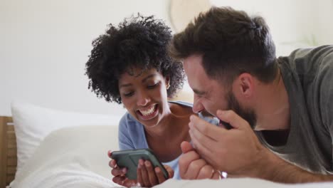 Video-De-Una-Pareja-Feliz-Y-Diversa-Relajándose-En-Casa,-Acostada-En-La-Cama-Usando-Un-Teléfono-Inteligente-Y-Riendo