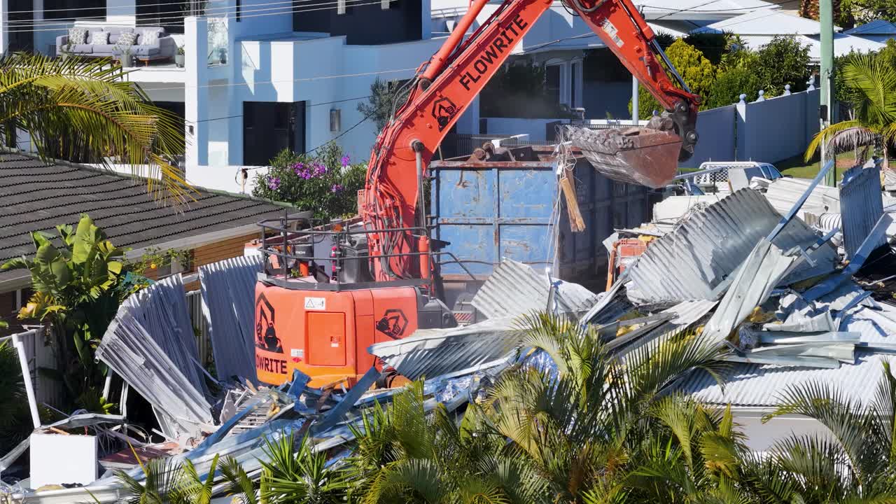Une pelleteuse orange démolit systématiquement une maison résidentielle au milieu d'une verdure luxuriante et de bâtiments modernes, filmée en plein jour avec un cadrage de caméra stable.