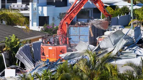 an orange excavator systematically demolishes a residential house amid lush greenery and modern buildings, captured in bright daylight with steady camera framing