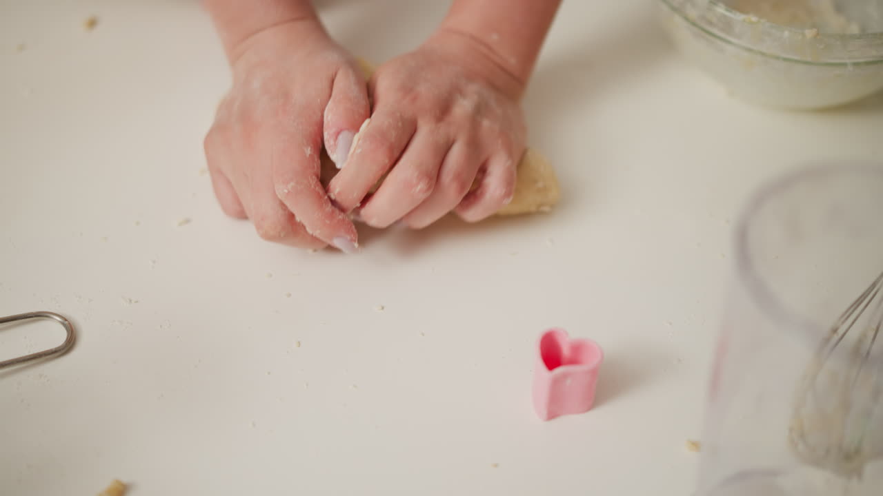 Hands moulding soft dough on floured table during baking process with person taking away roller pin, showing preparation stage with baking tools and pink heart shaped cookie cutter nearby