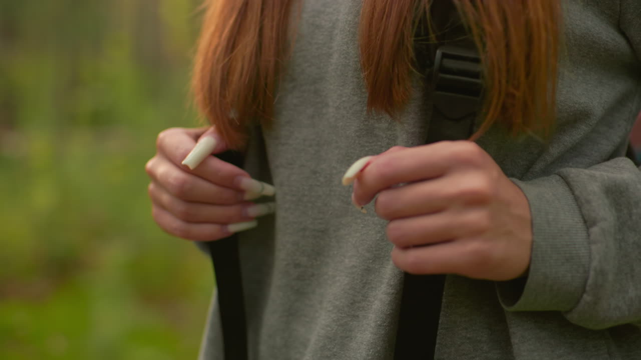 primer plano de una mujer caminando por un bosque exuberante, agarrando correas de mochila con uñas largas y cabello largo que fluye sobre los hombros, usando una sudadera gris y caminando a través de la vegetación