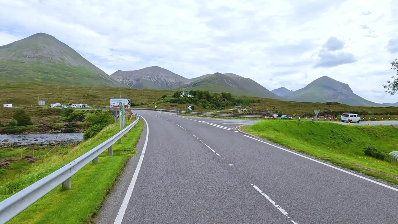 Forward-facing camera captures a smooth daytime drive along a winding rural road, surrounded by green hills and mountains under overcast skies