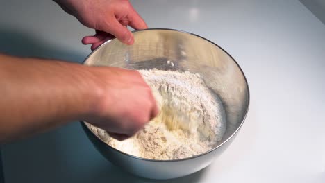 blending ingredients in a metallic mixing bowl placed on a white surface, dough for pizza and bread