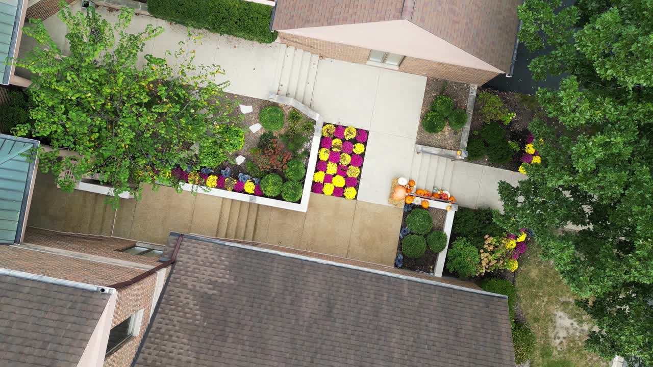 High-angle drone shot showing a residential courtyard featuring colorful fall flowers, pumpkins, and lush green trees between modern brick buildings