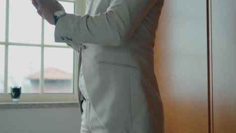 groom in elegant beige suit adjusting his watch near a window before the ceremony