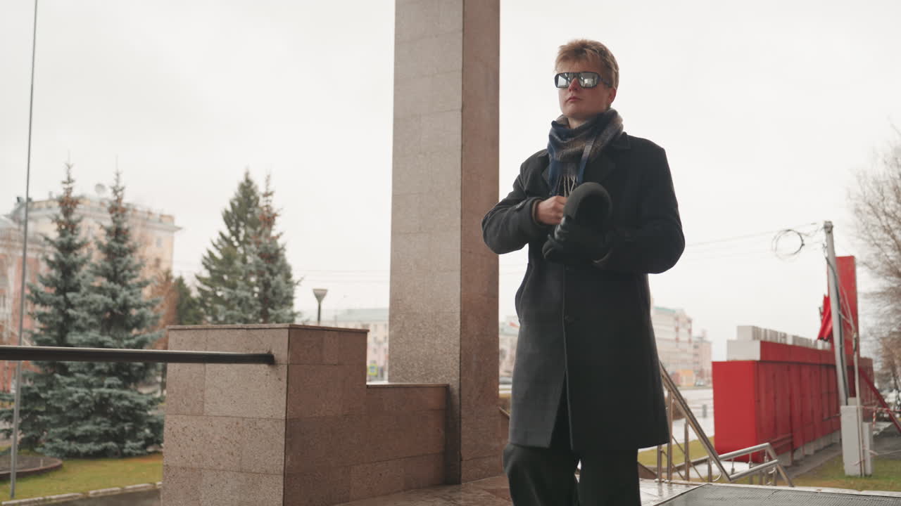 Porch urban creator white male filmmaker scouting location, wearing long coat and scarf, adjusting sunglasses, walking down steps, overcast sky, city trees and red kiosk in background, cinematic