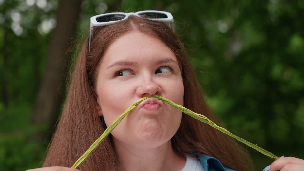 Close up of funny lady sitting outdoors with white sunglasses on head turning eyes sideways after using green thread to make playful pig face, surrounded by lush greenery