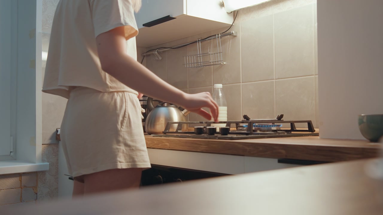 Scena serale in cucina, cuoco che regola la fiamma in una cucina compatta di appartamento con fornello a gas, movimento della mano concentrato, bollitore nelle vicinanze, luce soffusa, abbigliamento casual da casa e preparazione tranquilla del tè.