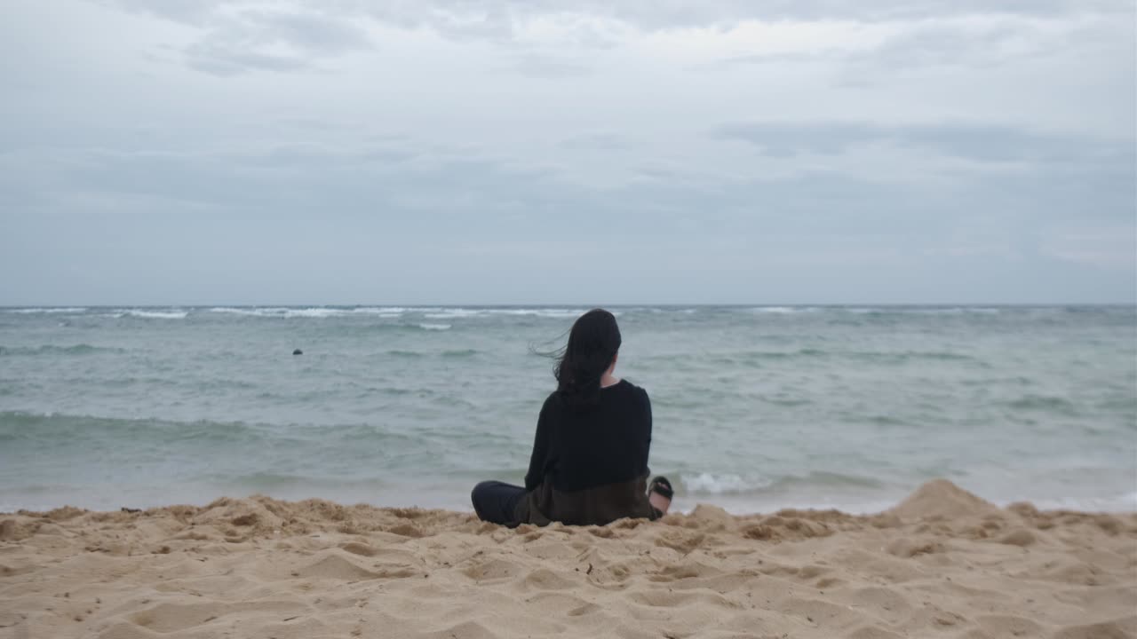 Rear view of young, woman sitting in deep thought at white sand beach with strong winds. -static shot